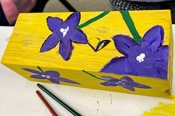 A decorated workshop bee hotel. It is painted bright yellow and has bluebell flowers painted on it.