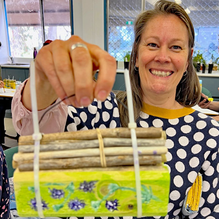 A participant at a community bee hotel workshop shows off her creation. The bee hotel is painted yellow, with purple flowers on green stalks.