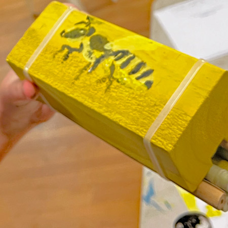 A completed bee hotel made during a workshop. It is painted light yellow and has a detailed image of a bee painted on the side.