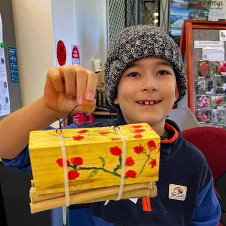 A school child participating in a bee hotel workshop proudly displays his finished work. The bee hotel is painted yellow with bright red flowers on green stalks.